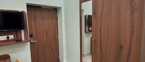 A room with a television mounted on the wall, a wooden wardrobe, a chair, and a tiled floor.