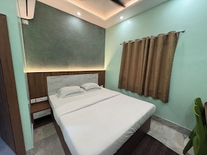 A neatly made bed with a headboard, a bedside table with a drawer, a wall-mounted air conditioner, and a curtain. - Hotel Sree Durga (Kolkata)