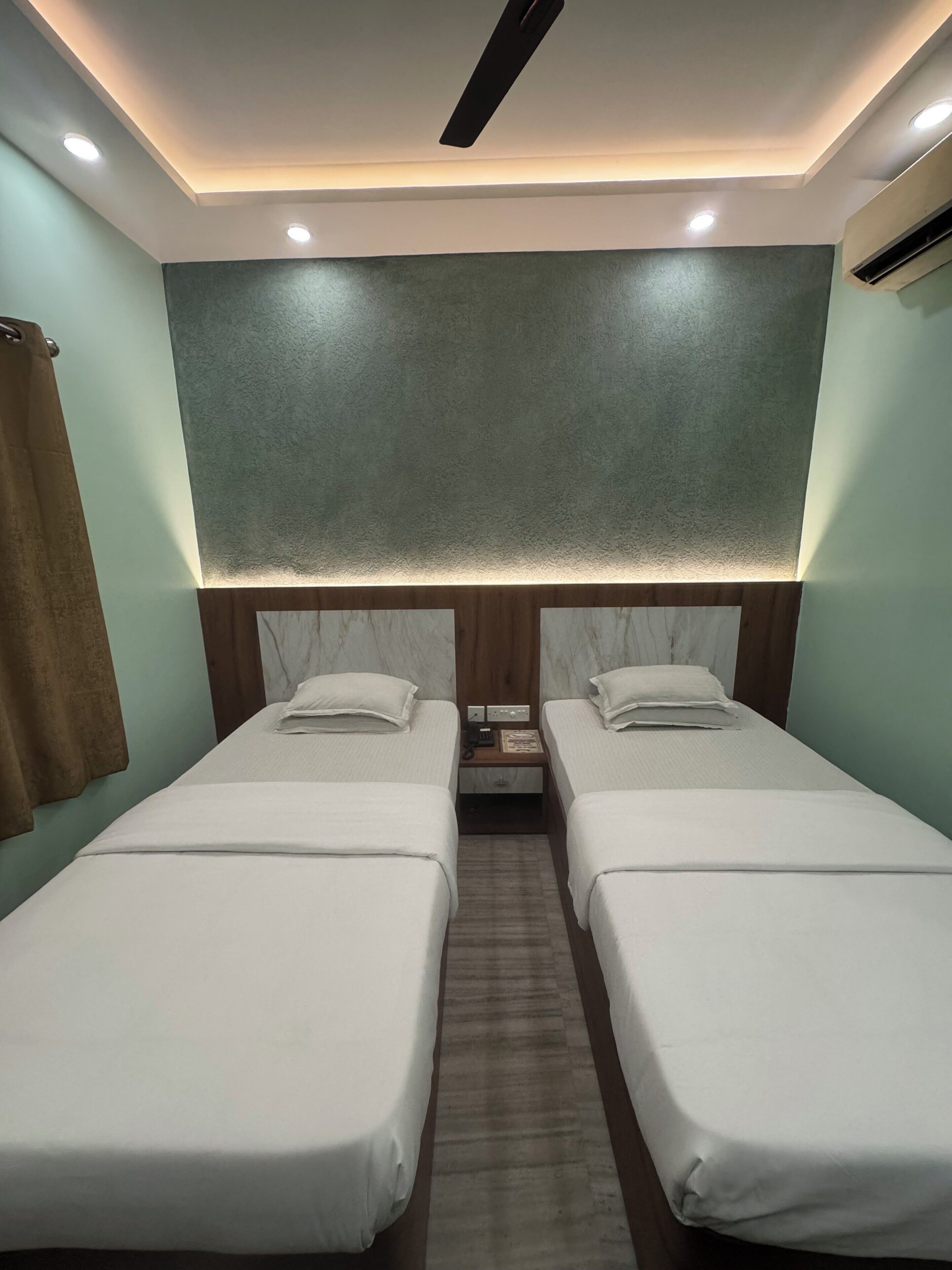 A hotel room with two single beds, a wooden headboard, a small bedside table, and a wall-mounted air conditioning unit.