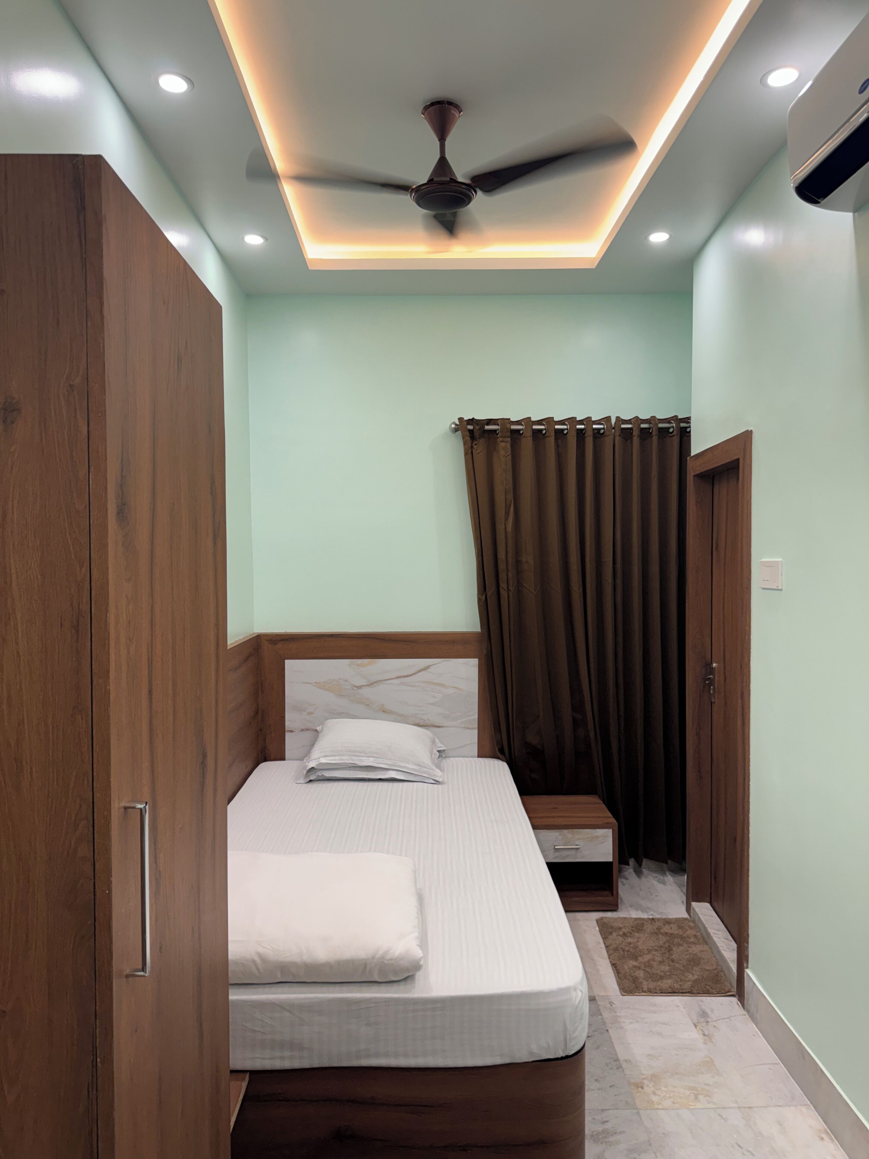 A bedroom with a bed, wooden wardrobe, ceiling fan, and a window with curtains.
