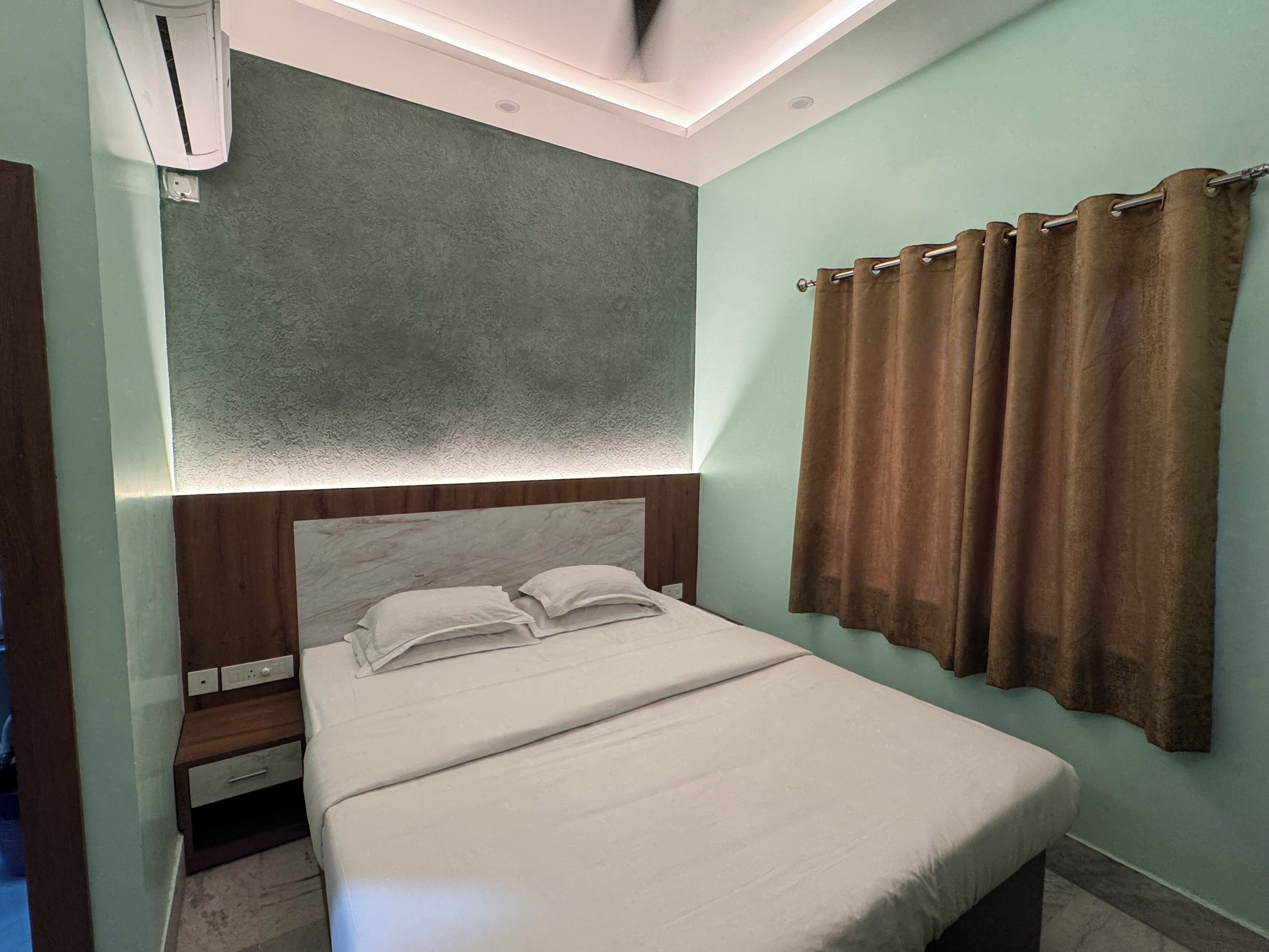 A neatly made bed with a headboard, a bedside table with a drawer, a wall-mounted air conditioner, and a curtain.