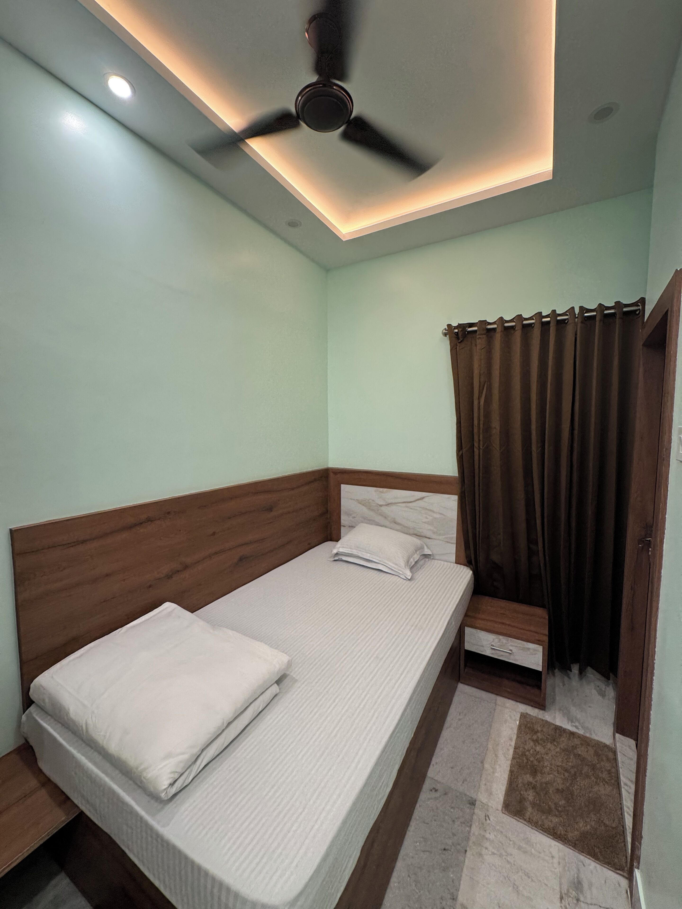A single bed with a wooden headboard, a ceiling fan, recessed lighting, and a brown curtain.