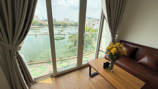 A room with a view of a river and buildings, a brown leather sofa, a wooden table, and a vase with yellow flowers. - Era Apartment Ho Rua (Hanoi)