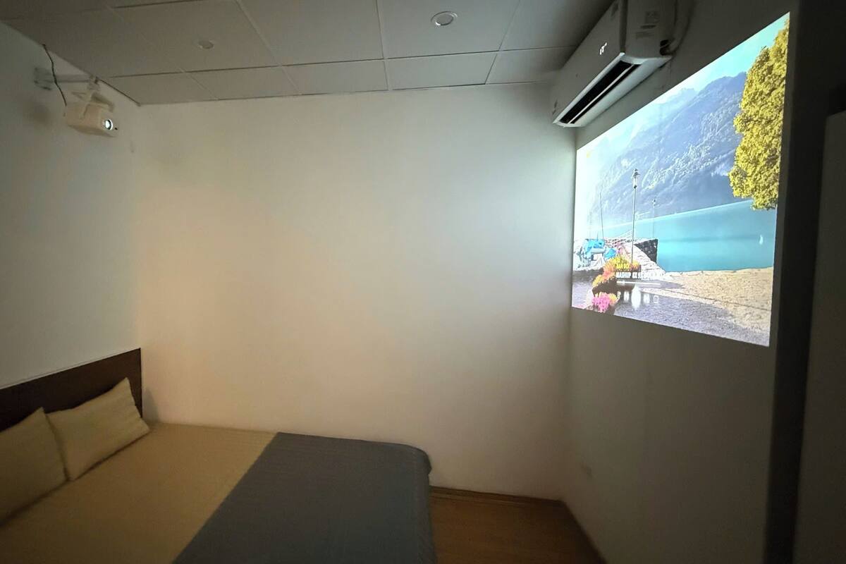 A small, simple room with a bed, a projector screen displaying a beach scene, and an air conditioning unit.