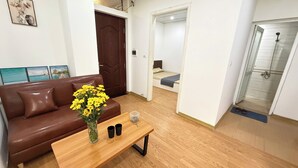 A compact living space with a brown leather sofa, a wooden coffee table with a vase of yellow flowers, a small table with two black cups, a wooden door, a bedroom with a bed and a shower area. - Era Apartment Ho Rua (Hanoi)