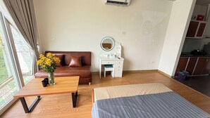 A modern bedroom with a wooden floor, a brown leather sofa, a white desk with a mirror, a bedside table with a vase of yellow flowers, and a large bed with a striped blanket. - Era Apartment Ho Rua (Hanoi)