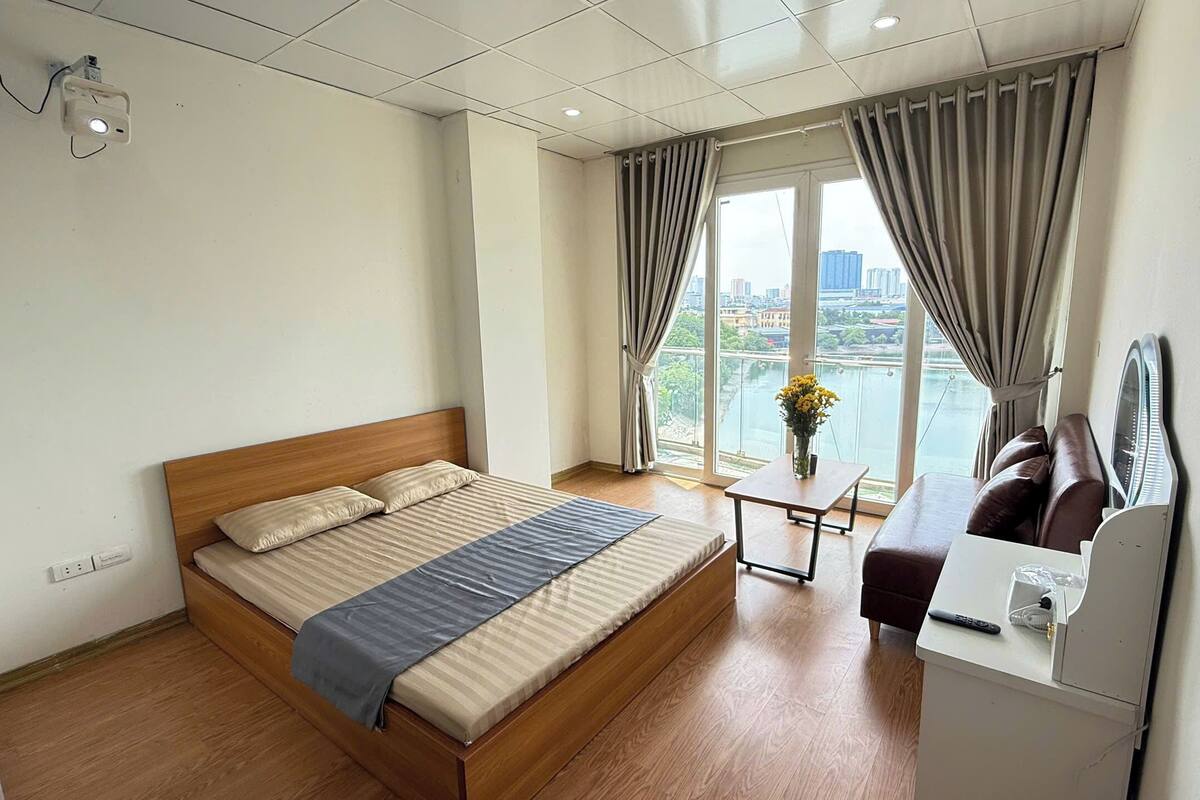 A hotel room with a wooden bed, a small table, a sofa, and a view of a cityscape through large windows.