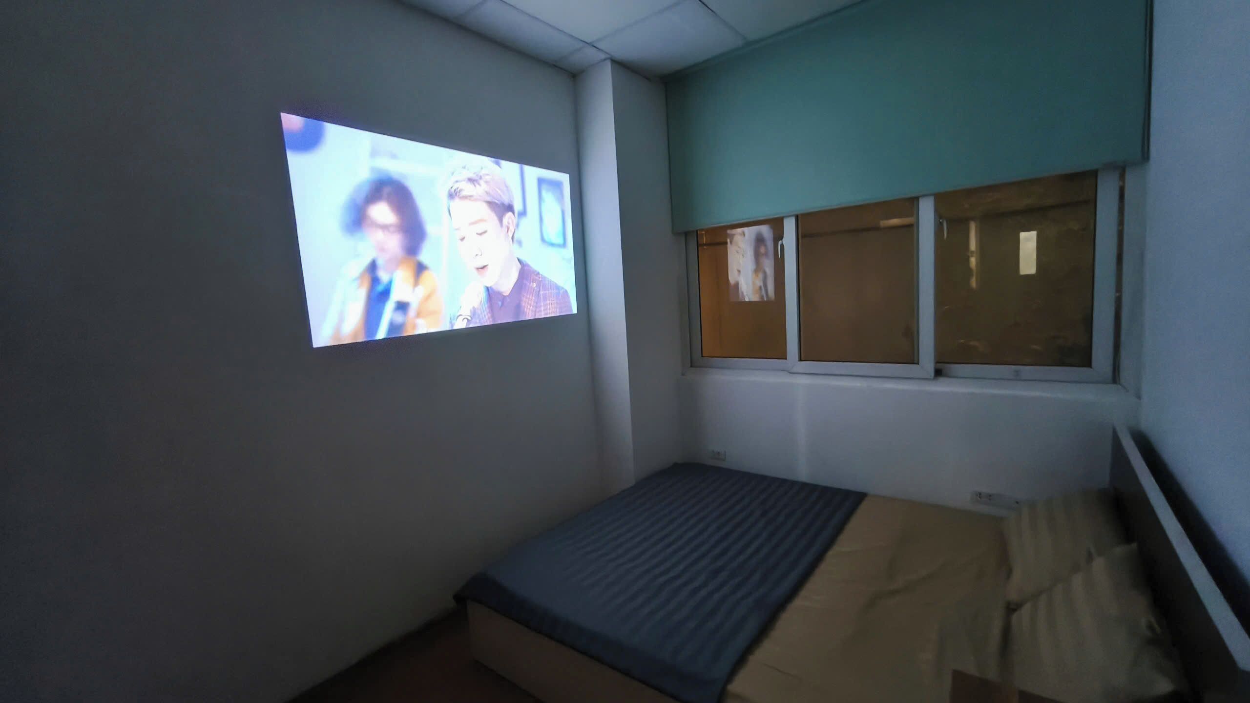 A small room with a bed, a window, and a projection screen displaying a scene.