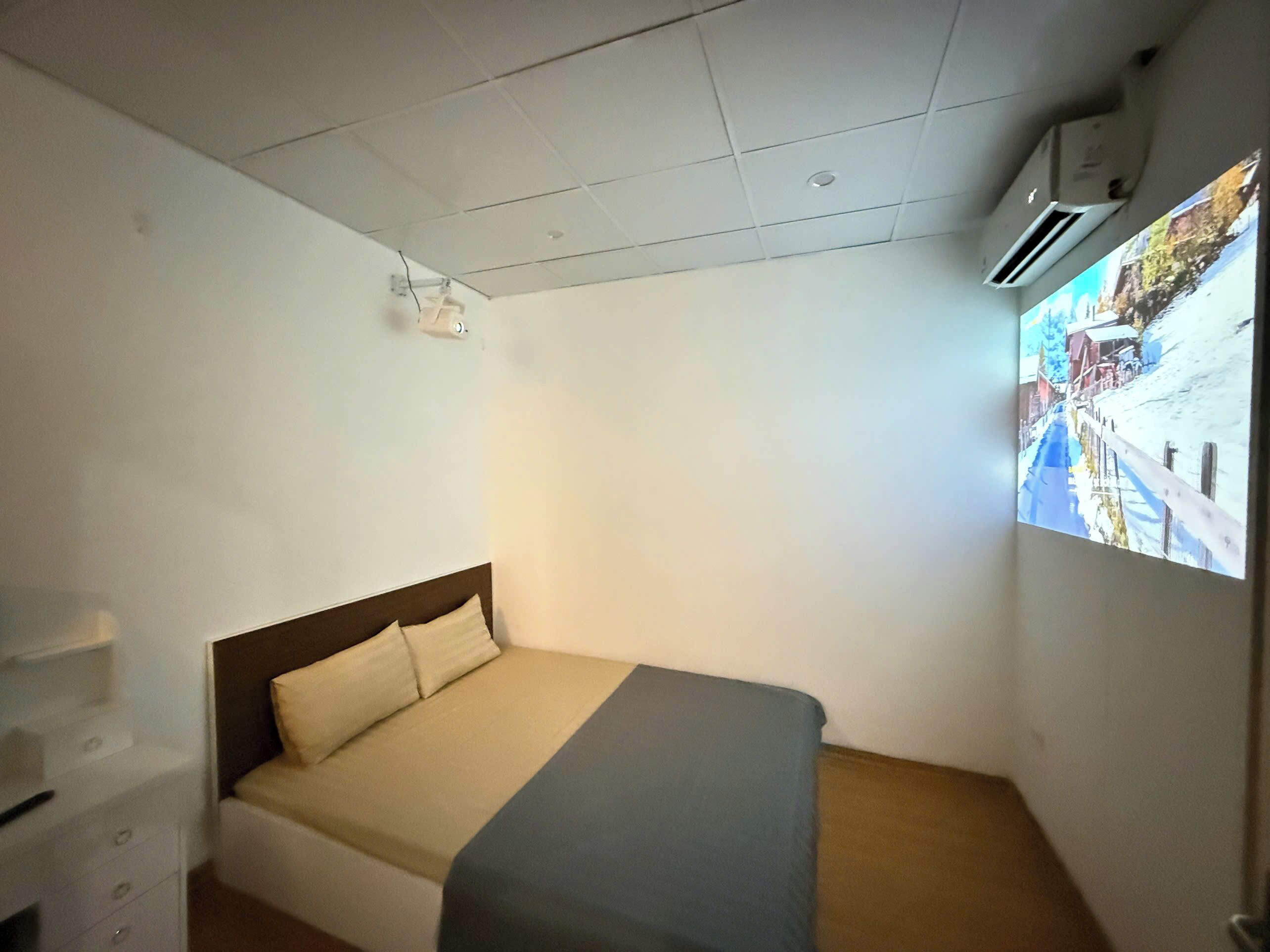 A small, single-bed room with a plain white wall, a wooden headboard, and a bed with a beige and gray blanket. There is a small shelf with a few items on it, and an air conditioning unit mounted on the wall.