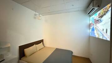 A small, single-bed room with a plain white wall, a wooden headboard, and a bed with a beige and gray blanket. There is a small shelf with a few items on it, and an air conditioning unit mounted on the wall.