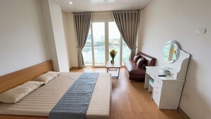 A hotel room with a bed, a sofa, a desk with a mirror, and a view of a body of water through large windows. - Era Apartment Ho Rua (Hanoi)