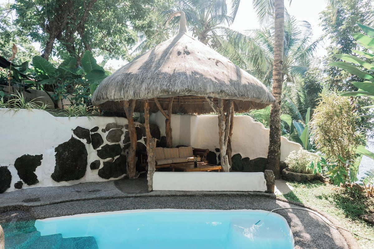 Estructura con techo de paja, sofá y mesa, piscina y vegetación tropical.