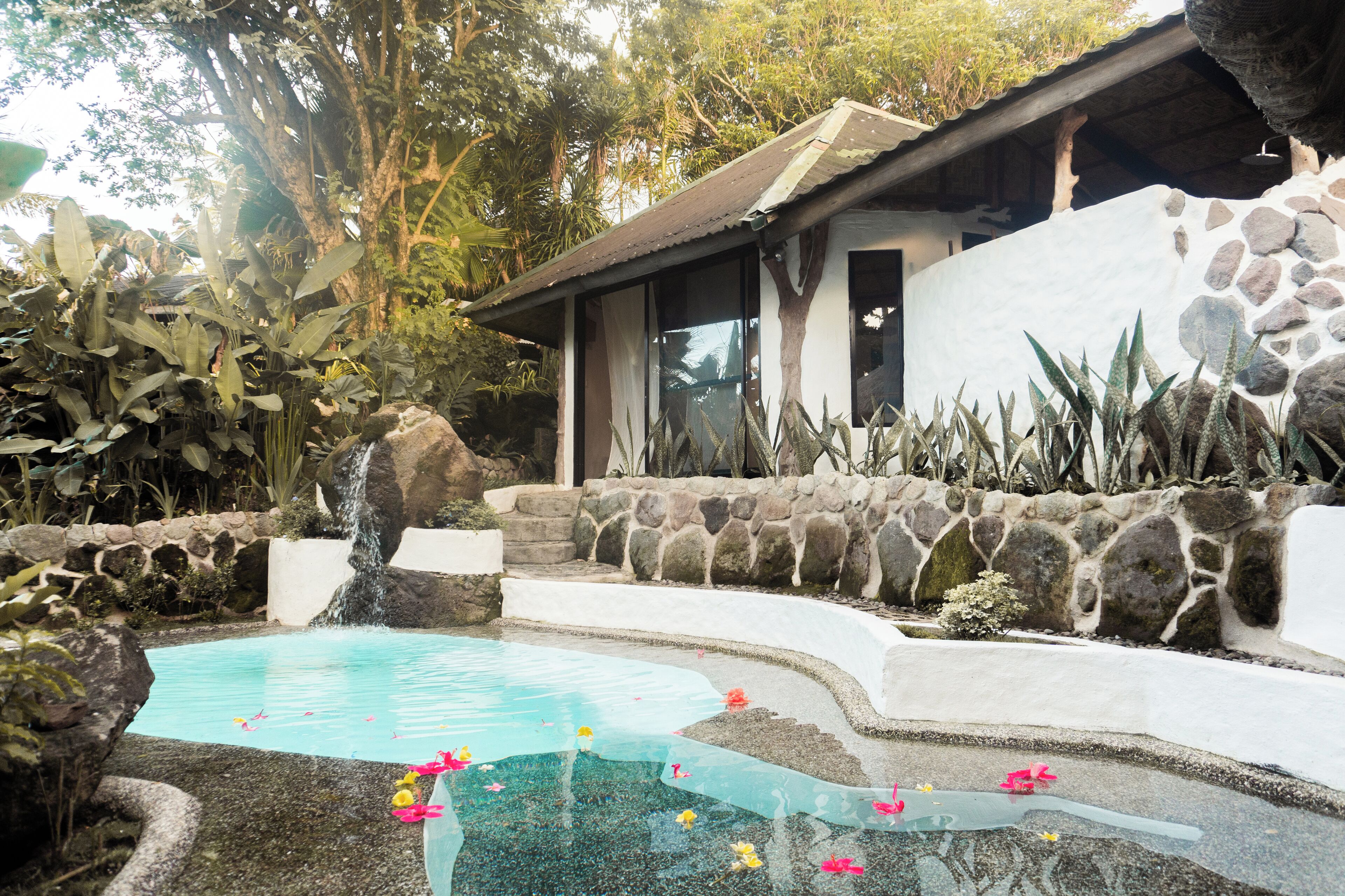 A pool with a waterfall, surrounded by tropical plants and a stone wall.