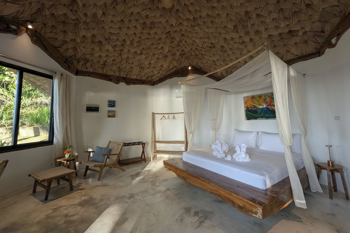 Un dormitorio con cama a dosel, un ventanal grande y un techo de materiales tejidos.