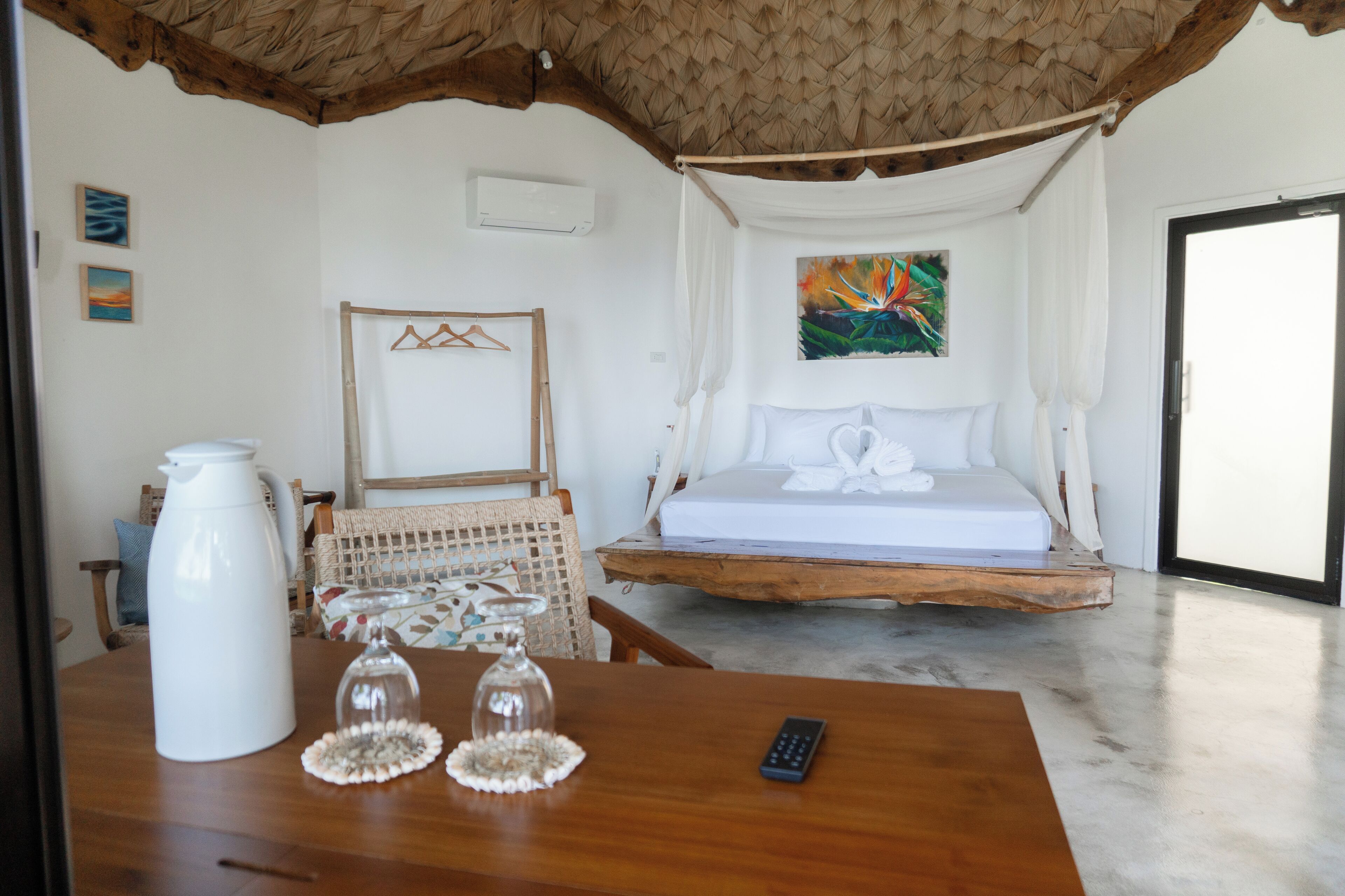 Un dormitorio con una cama, una mesa con una cafetera y vasos, una silla y un cuadro en la pared.
