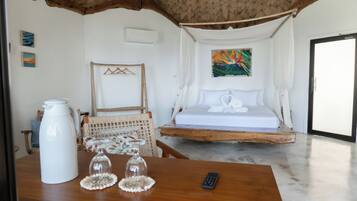 Un dormitorio con una cama, una mesa con una cafetera y vasos, una silla y un cuadro en la pared.