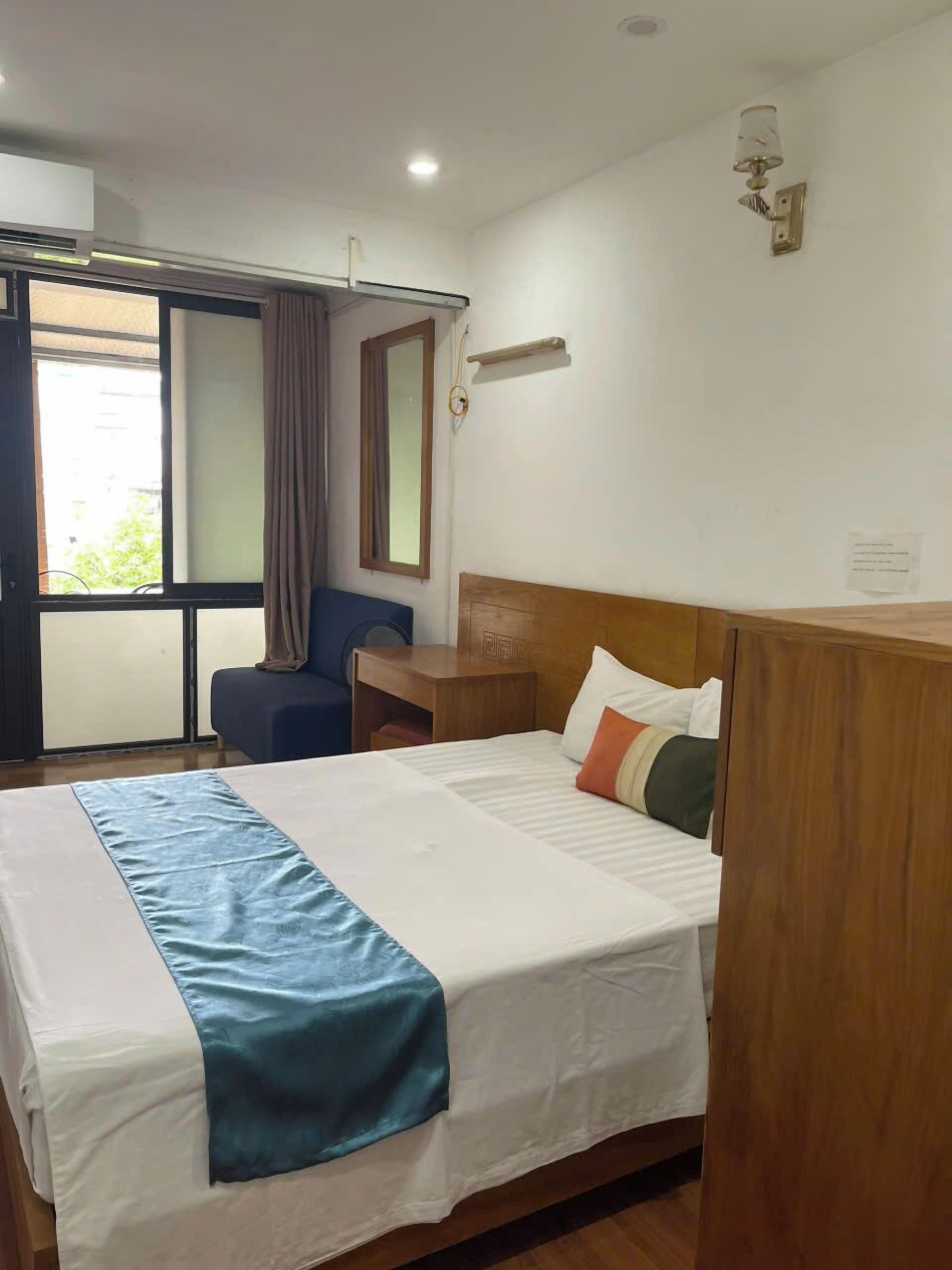 Ein Hotelzimmer mit Bett, Schreibtisch, Stuhl, einem Fenster mit Vorhängen und einer Wandlampe.