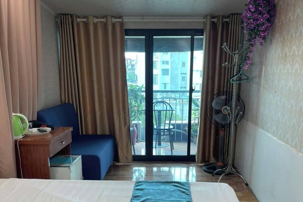 A hotel room with a bed, a desk, a chair, a balcony with a view of buildings, and an air conditioning unit.