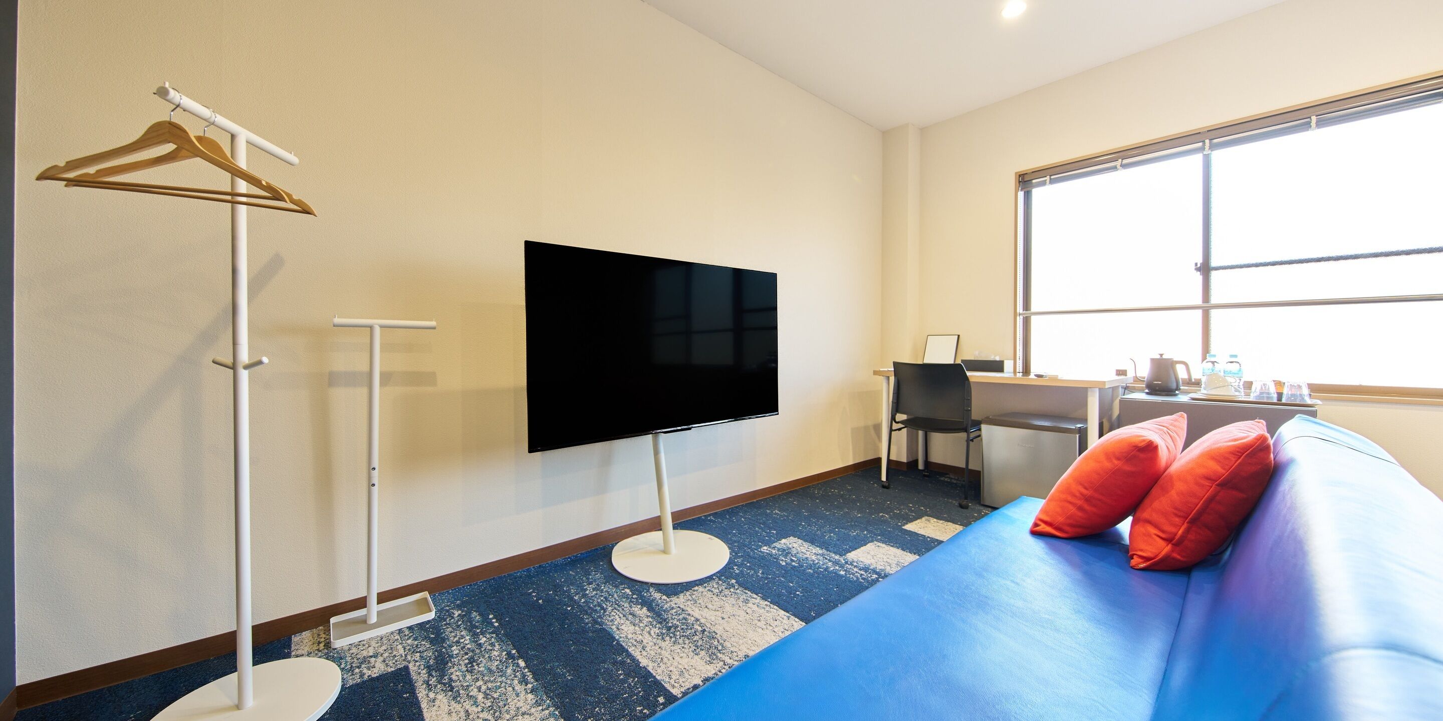 A hotel room with a large flat-screen TV mounted on the wall, a desk with a chair, a sofa with red cushions, and a large window providing natural light.