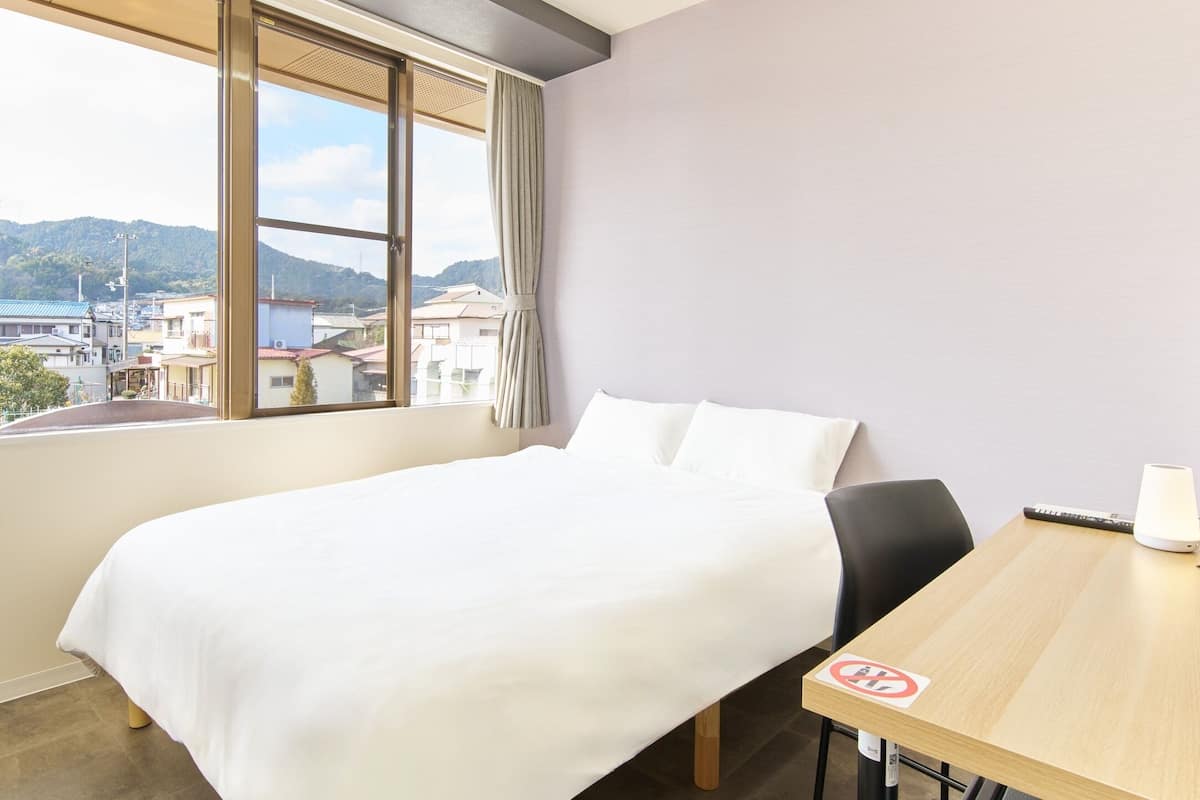 A hotel room with a bed, a desk with a lamp, and a view of a cityscape through large windows.
