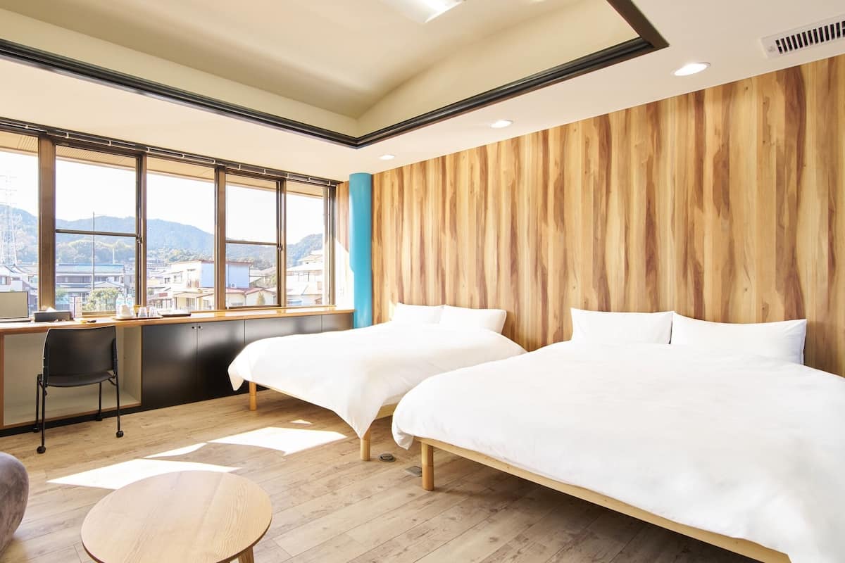 A hotel room with two beds, a desk, a chair, and a view of a cityscape.