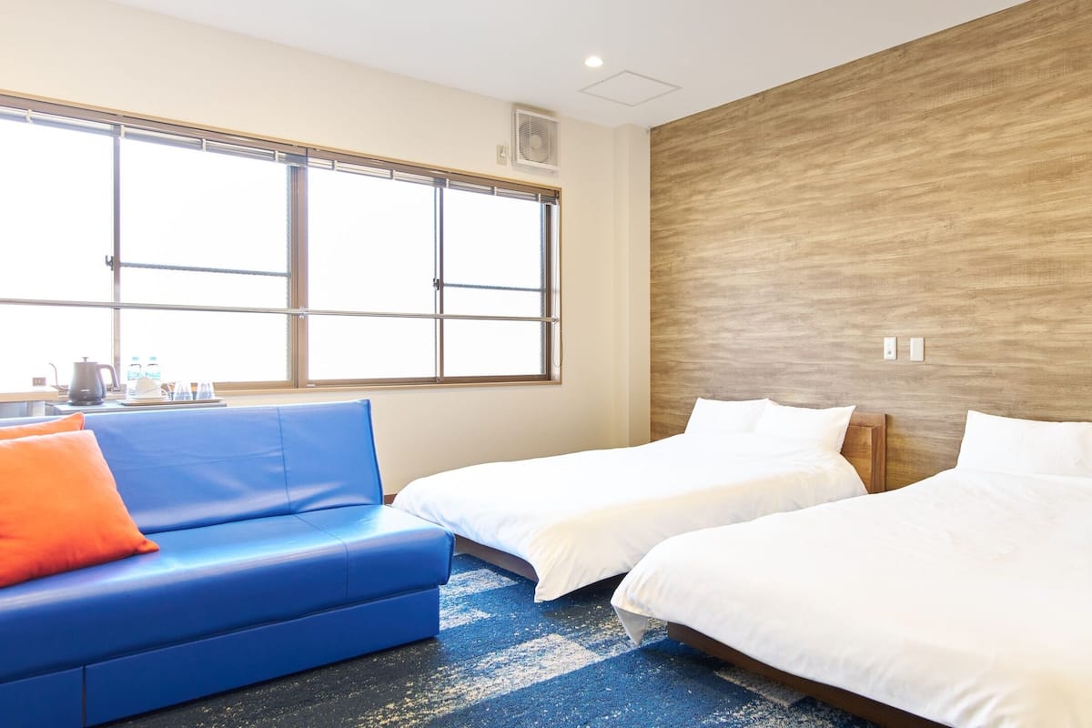 A hotel room with a blue sofa, two beds, a large window, and a wooden accent wall.