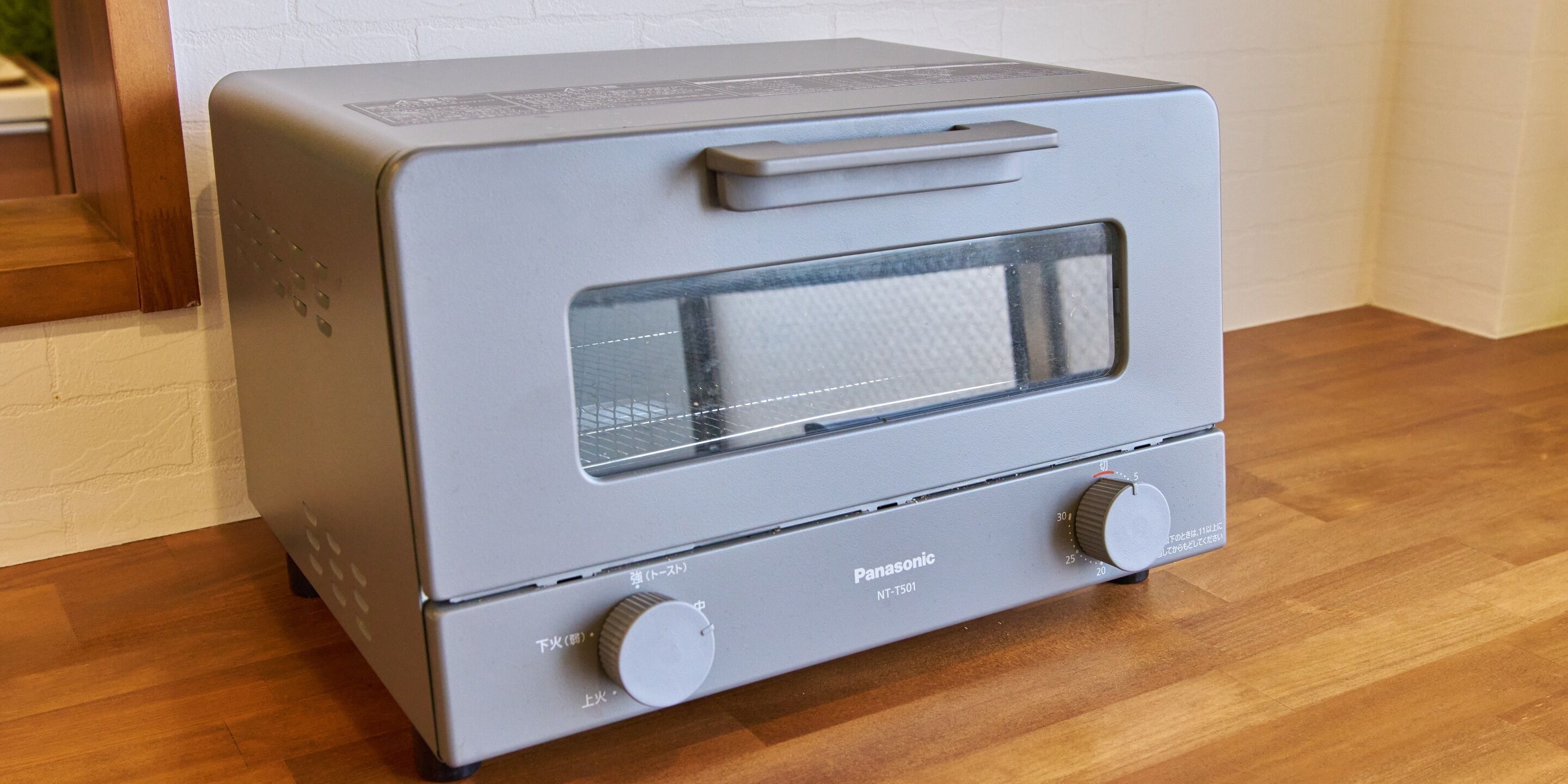 A Panasonic NT-T501 toaster oven with a handle and two knobs, placed on a wooden floor against a white brick wall.