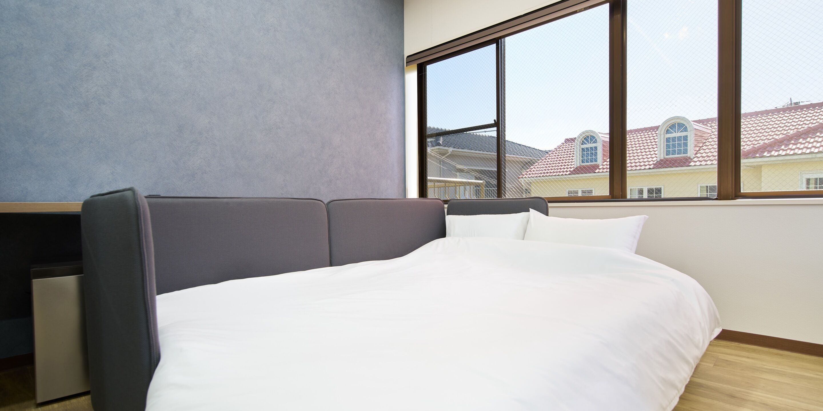 A modern bedroom with a large window, a bed with white bedding, and a grey headboard.