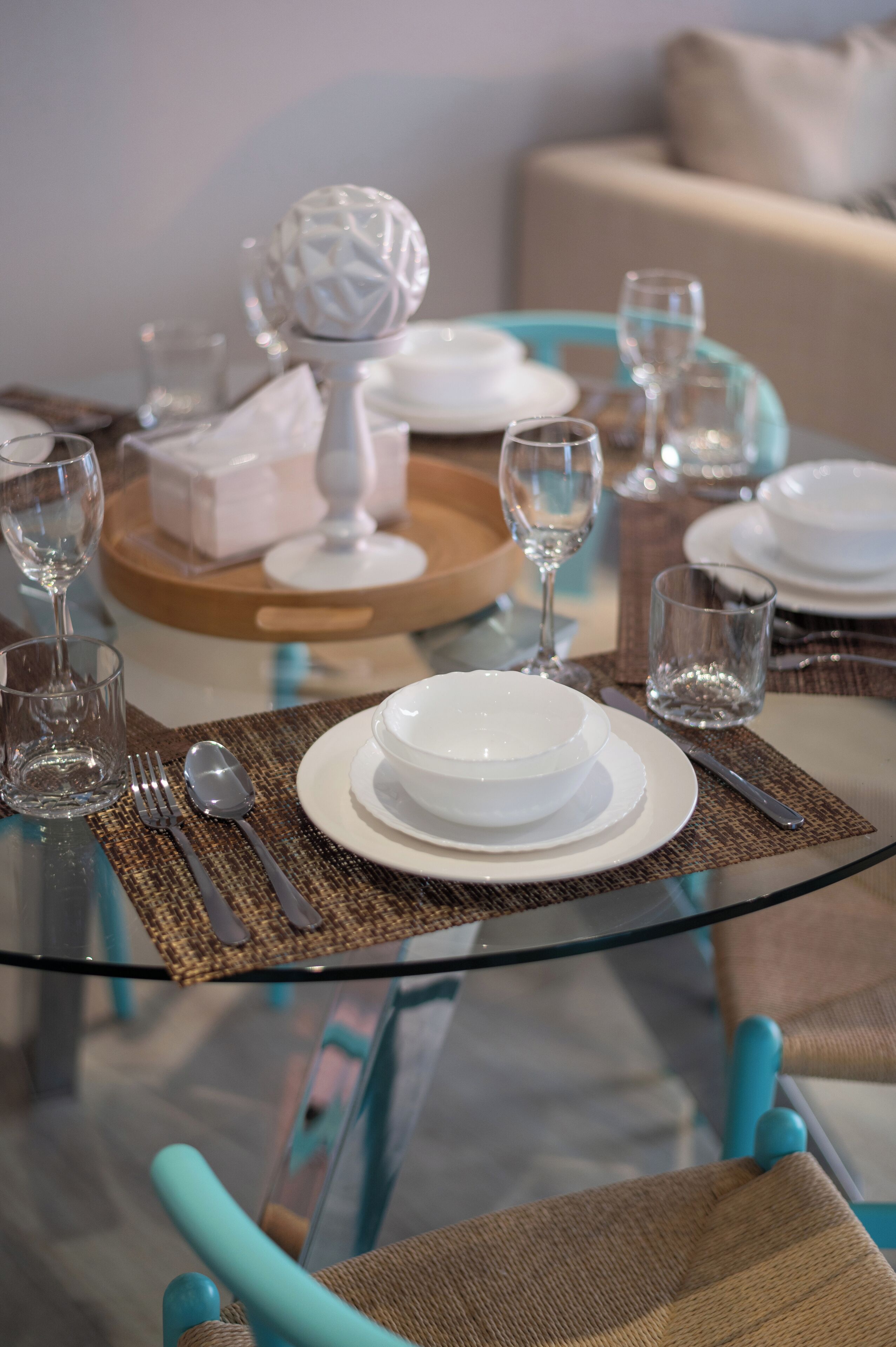 A dining table set with white plates, bowls, and cutlery, a glass vase, and a tray with a white object.