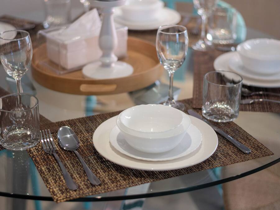 A dining table set with white plates, bowls, and cutlery, a glass vase, and a tray with a white object.