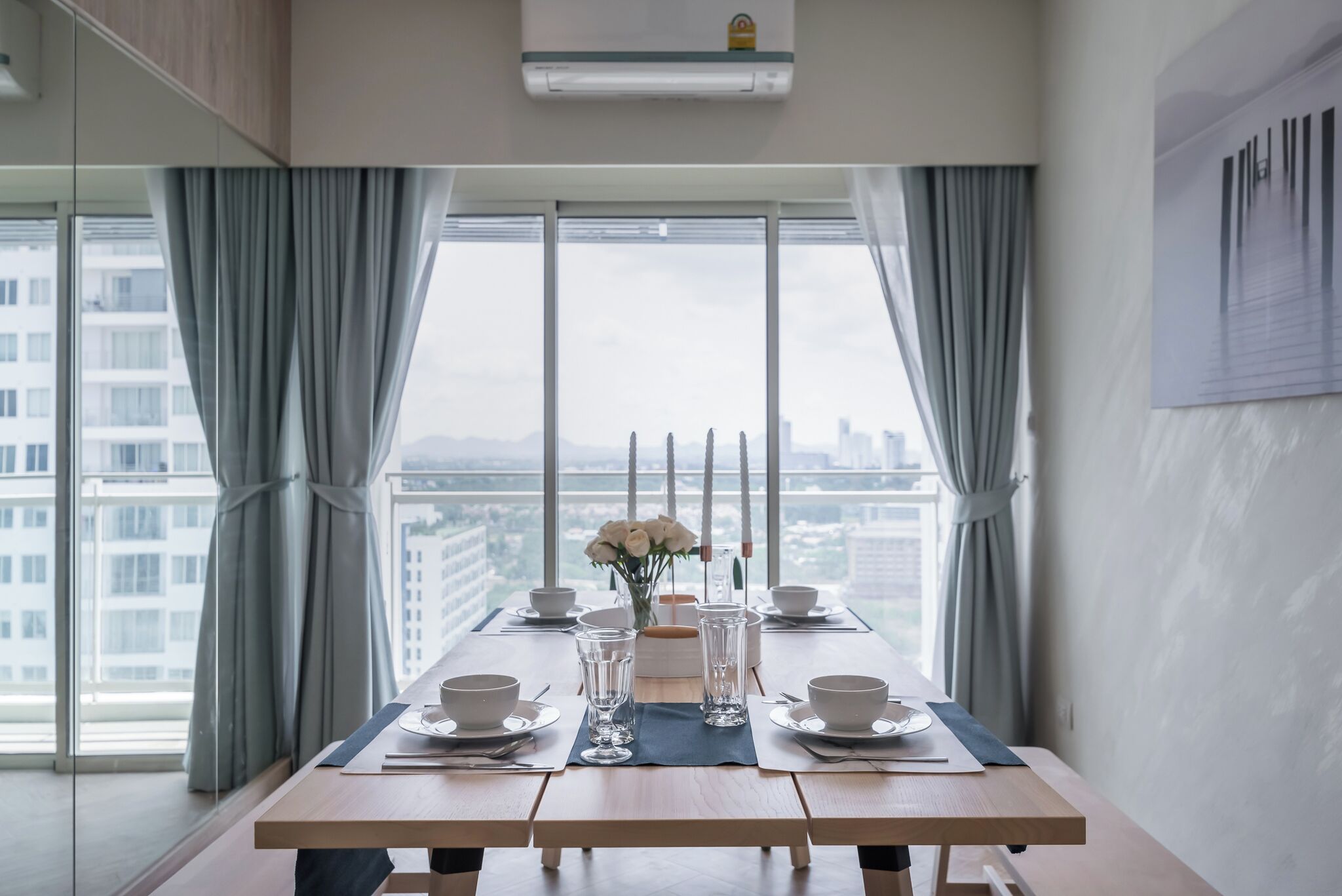 A dining table set for four with a vase of white flowers, a candle, and a view of a cityscape through large windows.