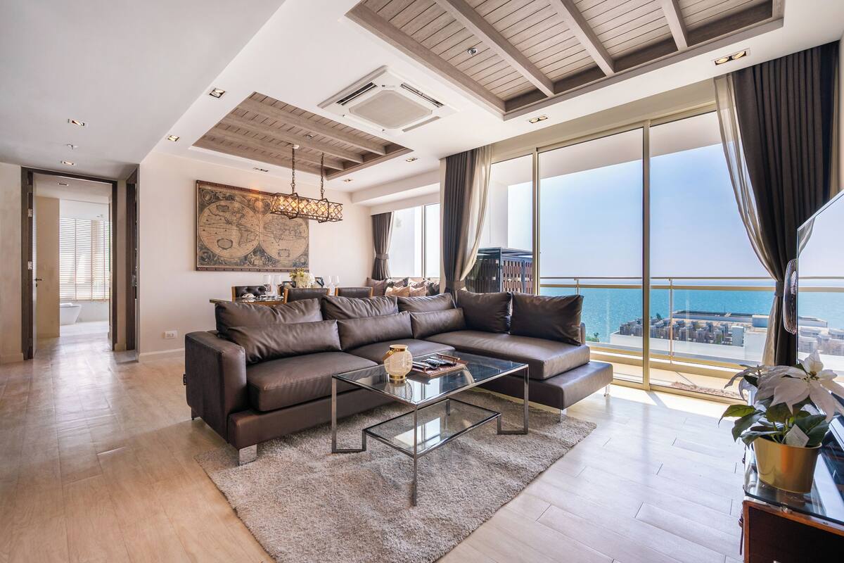 A modern living room with a large sectional sofa, a glass coffee table, and a large window offering a view of the sea.