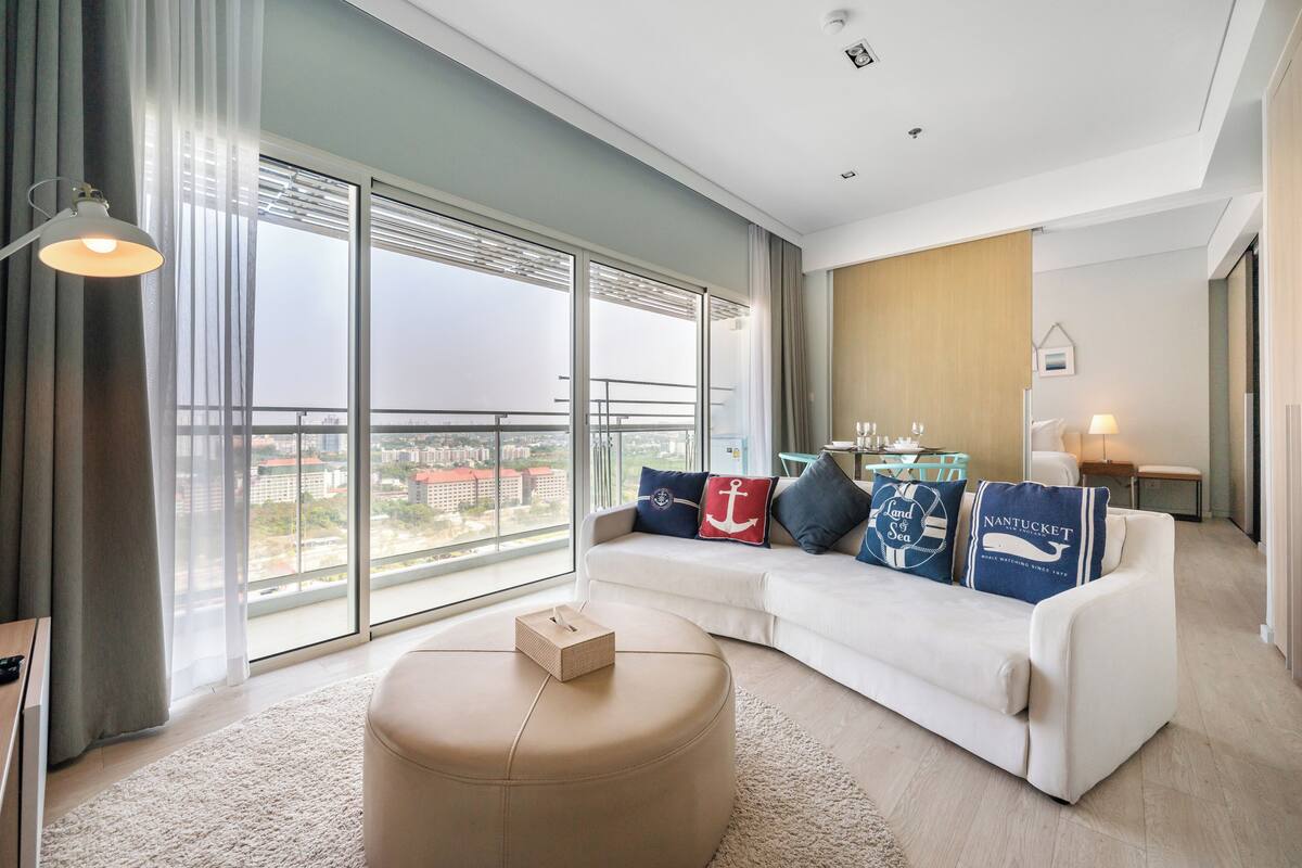 A modern hotel room with a large balcony, a white sofa with nautical-themed pillows, a round ottoman, and a view of the cityscape.