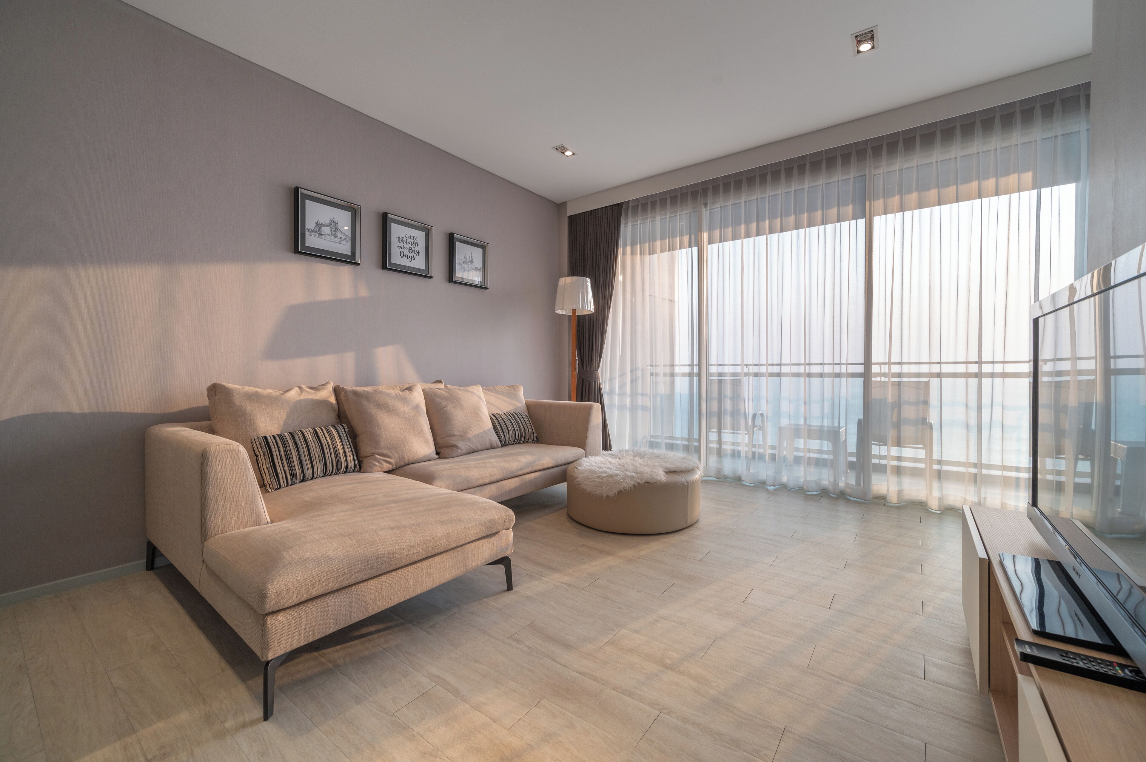 A modern living room with a beige sofa, a round ottoman, and a flat-screen TV. There are three framed pictures on the wall and a large window with sheer curtains.
