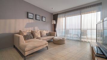 A modern living room with a beige sofa, a round ottoman, and a flat-screen TV. There are three framed pictures on the wall and a large window with sheer curtains.