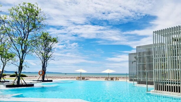 A clear blue pool with lounge chairs and umbrellas, surrounded by trees and a view of the beach. - Veranda Residence by SEA U PATTAYA (Pattaya)
