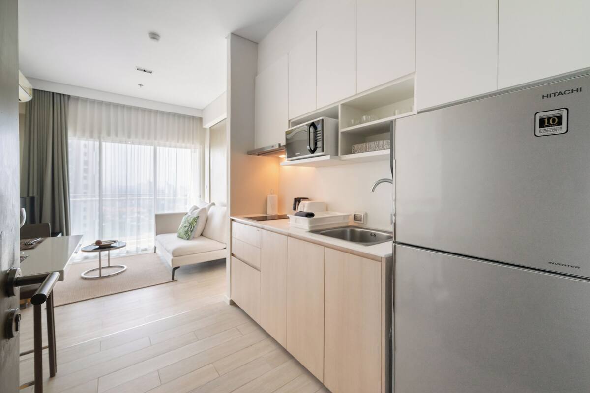 A modern kitchen with a stainless steel refrigerator, a microwave, and a sink. There is a dining area with a table and chairs, and a sofa in the background.