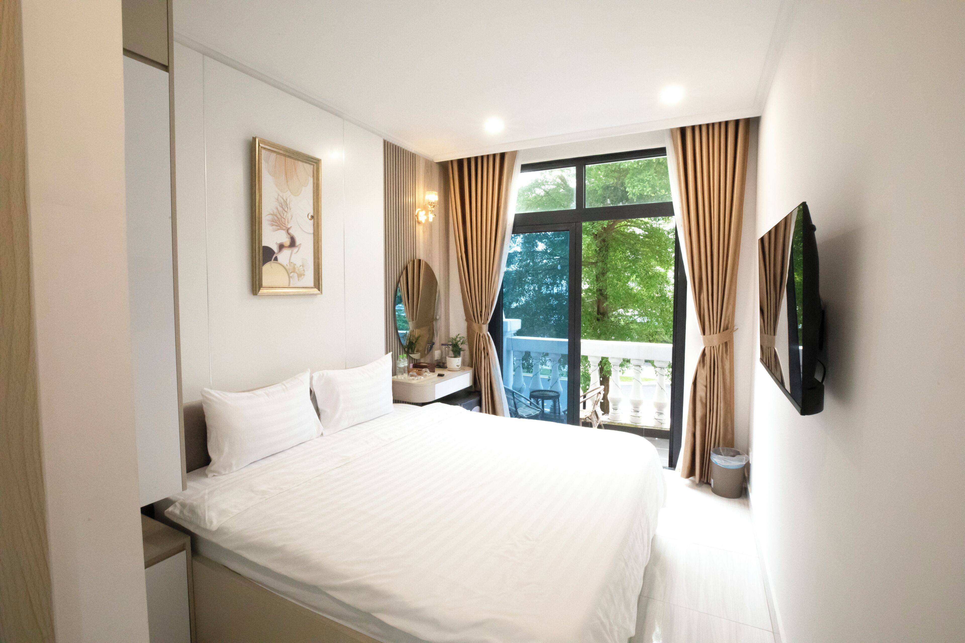 A bedroom with a large bed, a bedside table with a lamp, a window with curtains, and a wall-mounted TV.