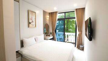 A bedroom with a large bed, a bedside table with a lamp, a window with curtains, and a wall-mounted TV.