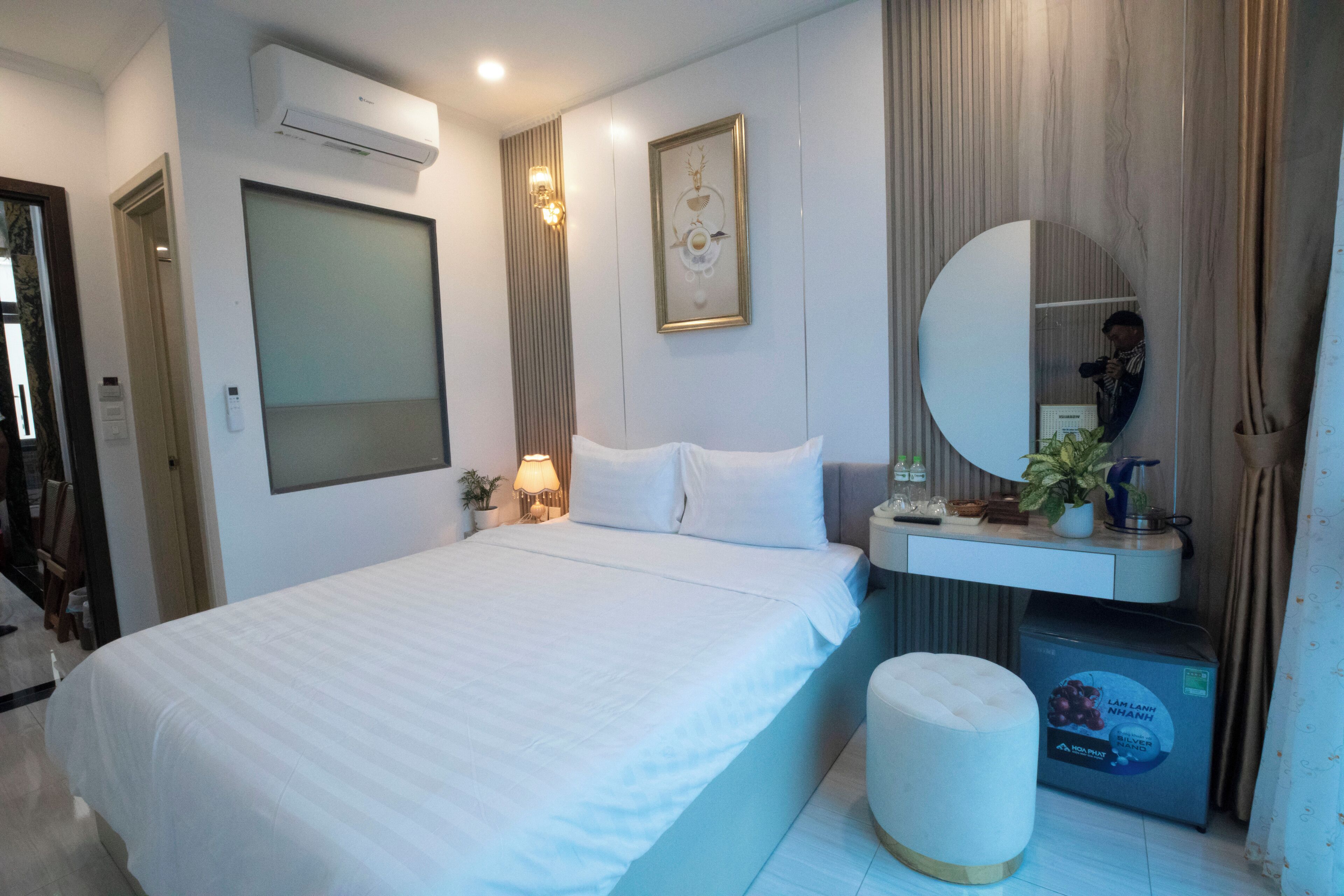 Een moderne hotelkamer met een groot bed, een nachtkastje met een lamp, een spiegel, een kleine potplant en een minibar.