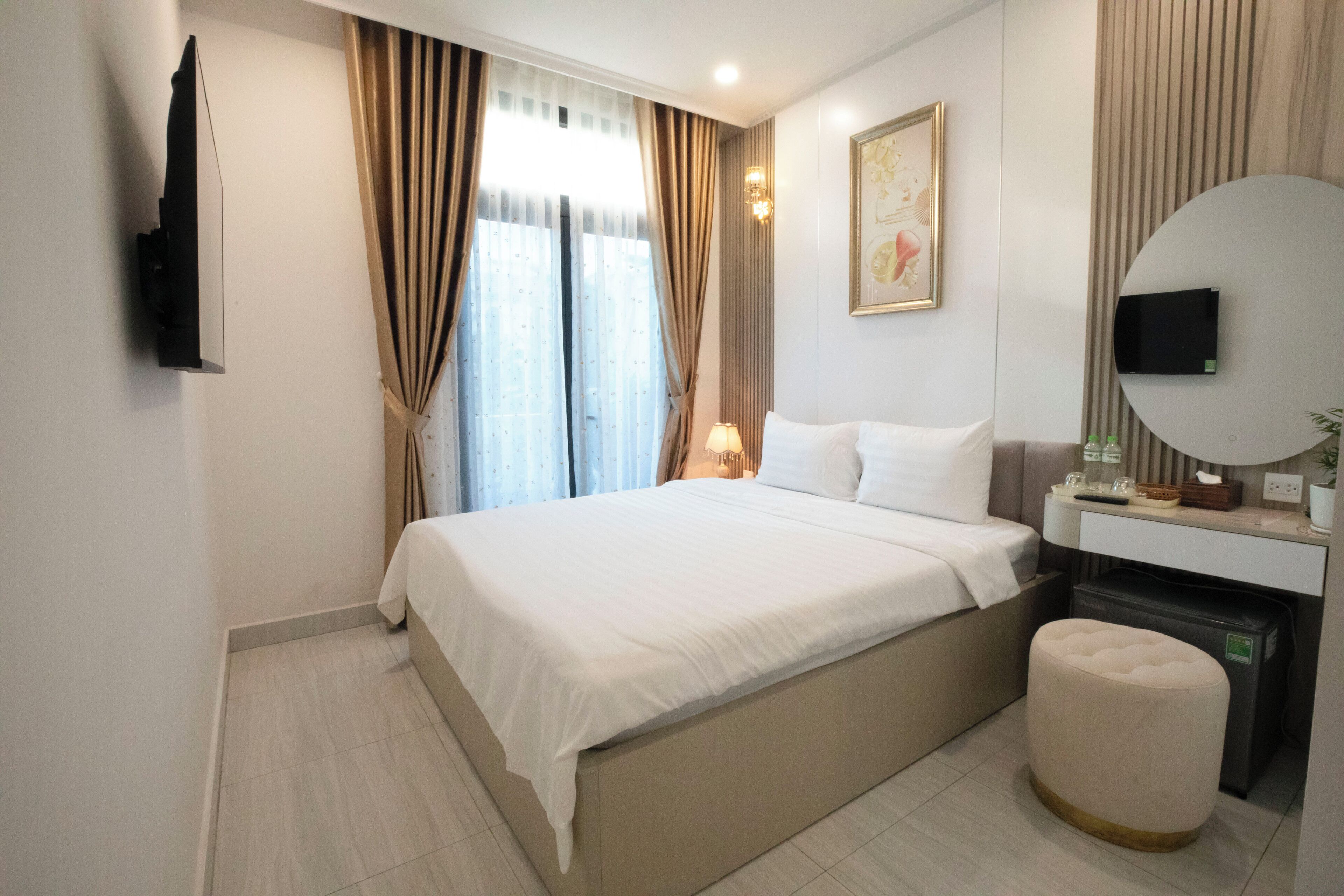 A modern hotel room with a large bed, a bedside table with a lamp, a television mounted on the wall, and a small stool.