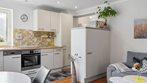 A modern kitchen with white cabinets, a built-in oven, and a dishwasher. There is a dining area with a round table and chairs, and a gray sofa with a small white table beside it. - Holiday apartment near the beach with a terrace (Hohenkirchen OT Wahrstorf)