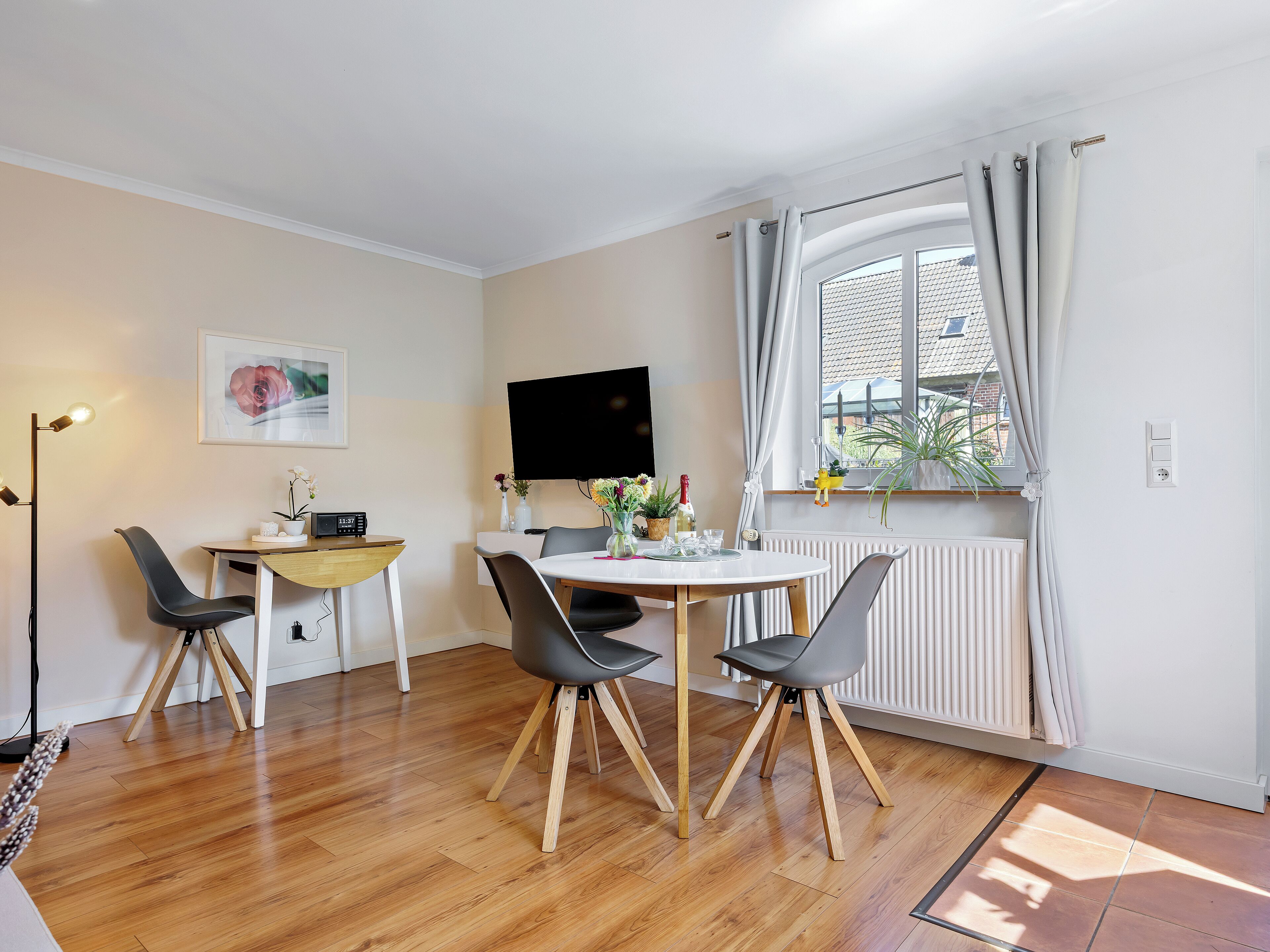 A modern living room with a dining area, wooden flooring, a flat-screen TV, and a window with curtains.