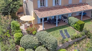 Une maison à deux étages avec un toit en tuiles, un patio couvert avec des meubles d’extérieur et une pelouse agrémentée de deux chaises longues.