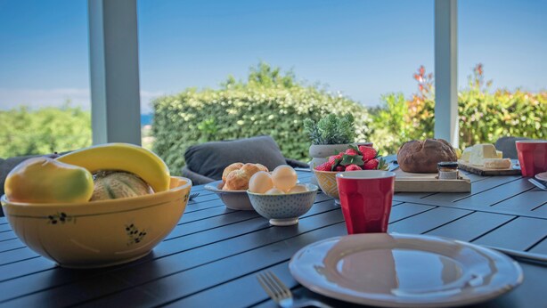 Une table à manger, dressée avec divers plats et couverts, se détachant sur un fond de verdure et d’un ciel dégagé.