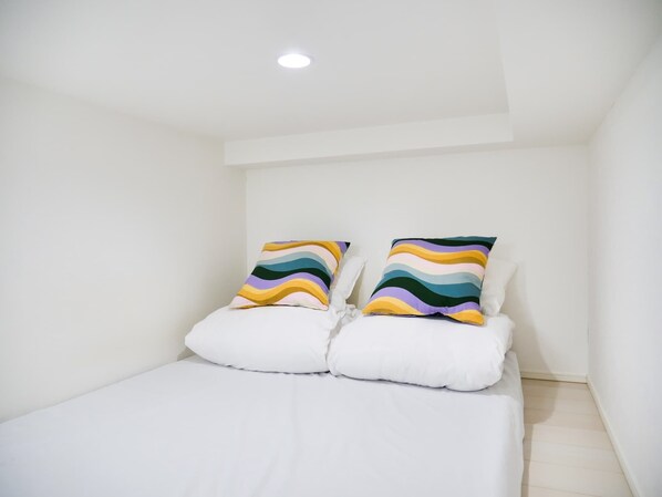 A small, white, single-bed room with two colorful pillows and a white bedspread.