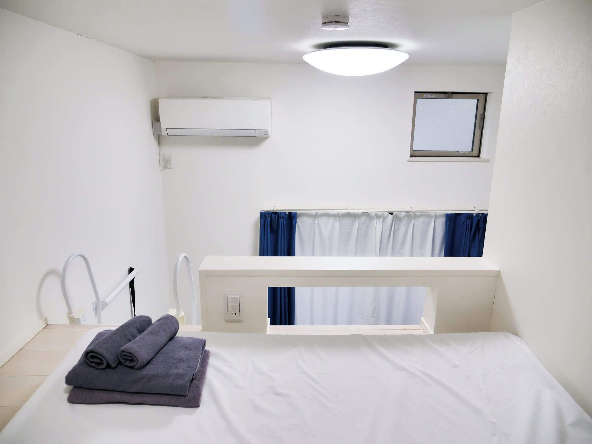 A hotel room with a bed, a white air conditioning unit, a window with blue curtains, and a small mirror on the wall.