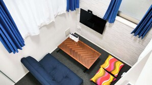 A compact room with a sofa, a small wooden table, a TV mounted on the wall, and a window with curtains. - Triphome Koenji Minami 4 mins Higashi Koenji stn (Suginami City)