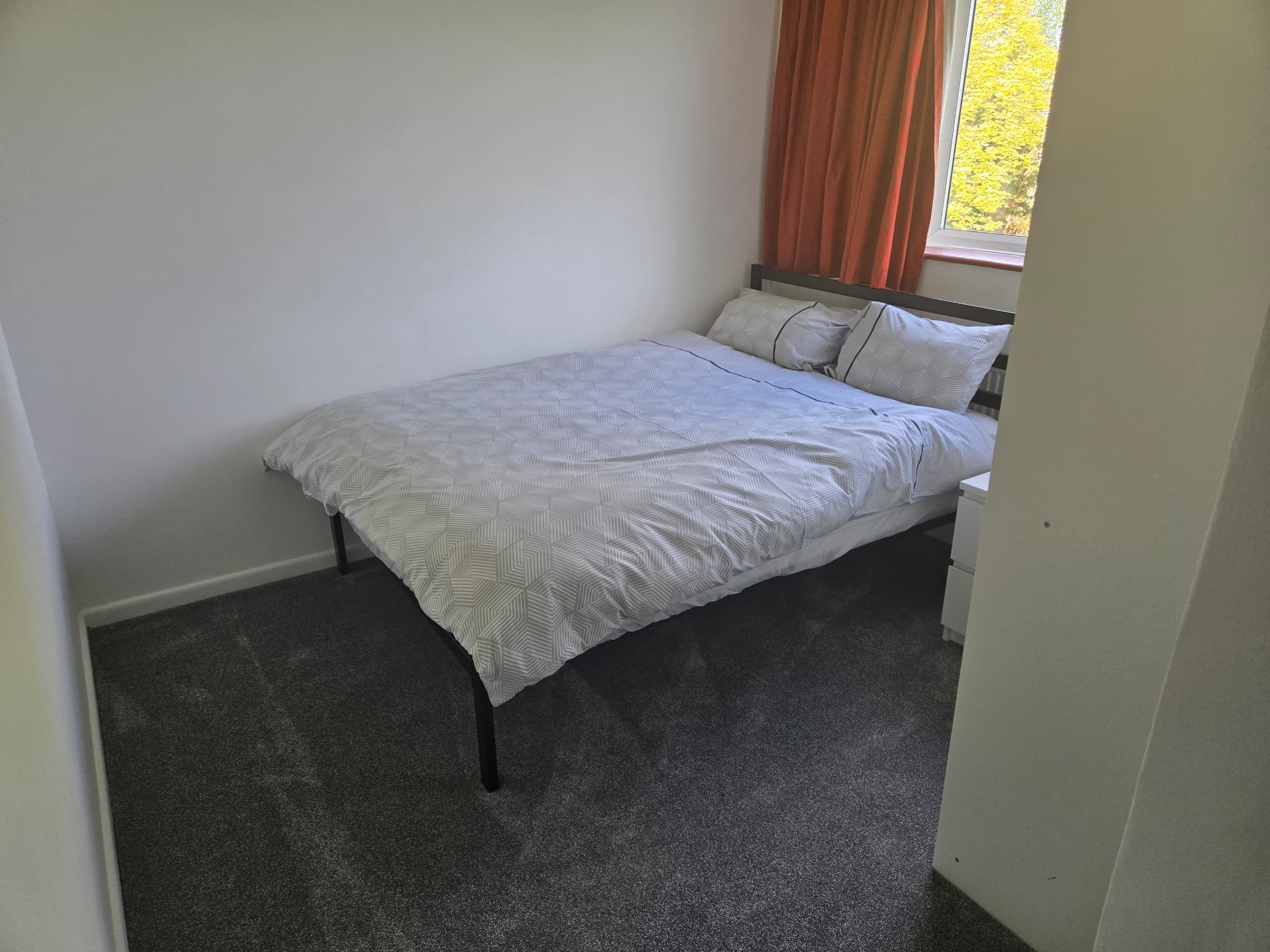 A single bed with a patterned bedspread, a window with orange curtains, and a plain carpeted floor.