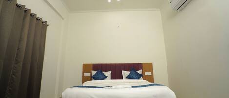 A hotel room with a bed, curtains, an air conditioning unit, and a ceiling light.